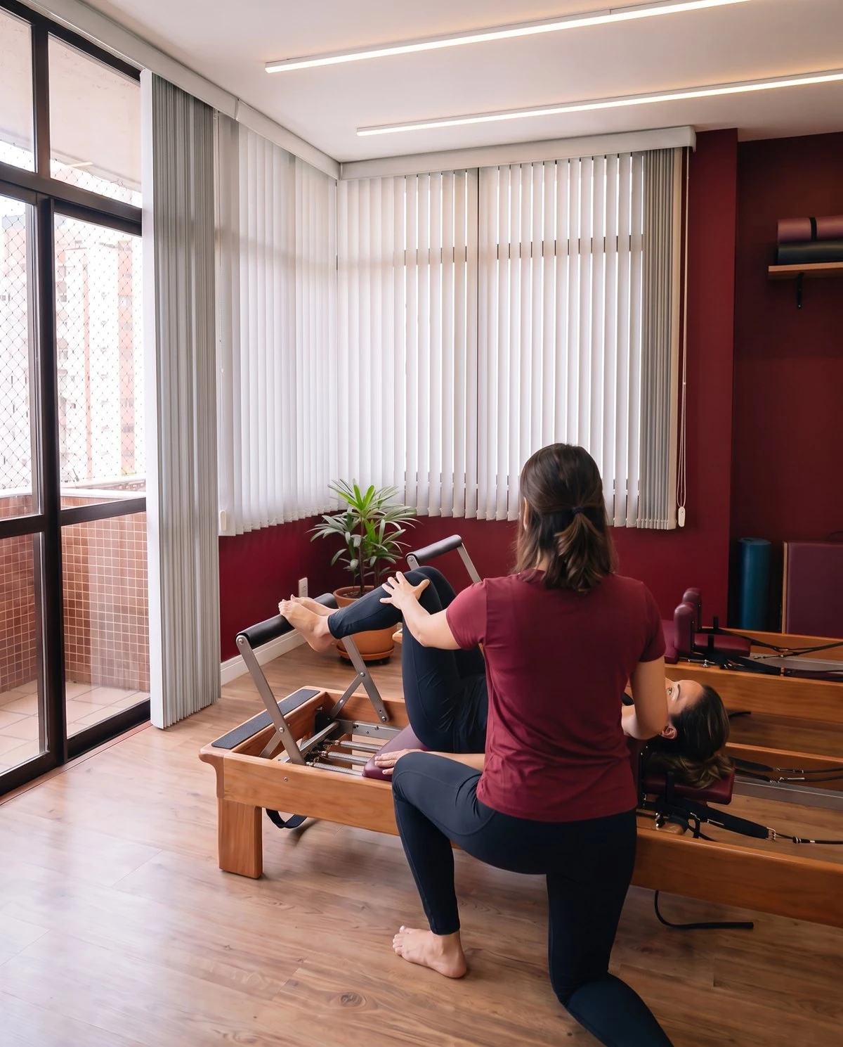 Instrutora de pilates, de costas, orientando uma aluna deitada no Reformer, com a parede bordô ao fundo