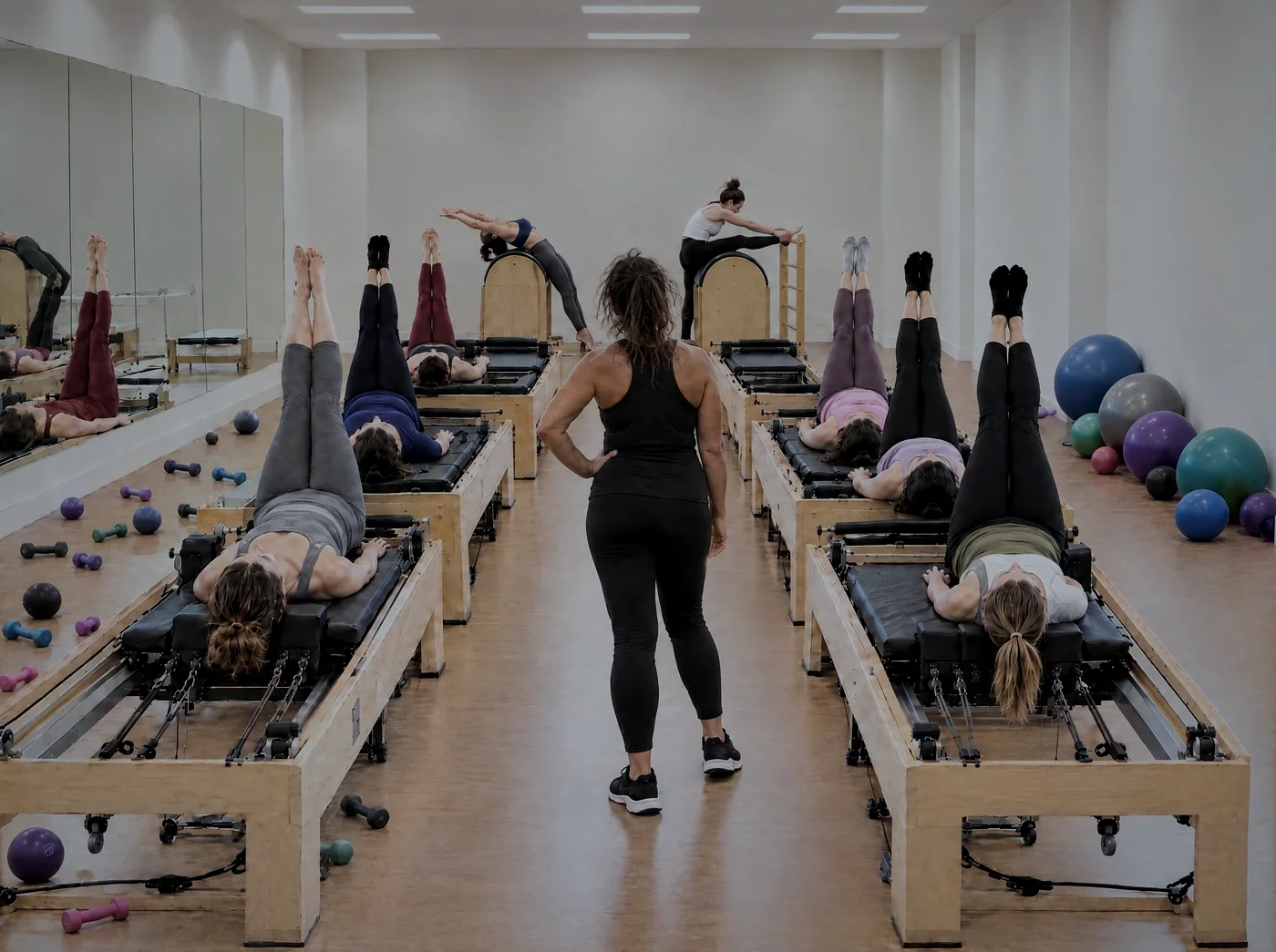 Aula de pilates em grupo, com duas fileiras de reformers ocupados, duas alunas em Ladder Barrels ao fundo e uma instrutora de costas no corredor, marcada com um X